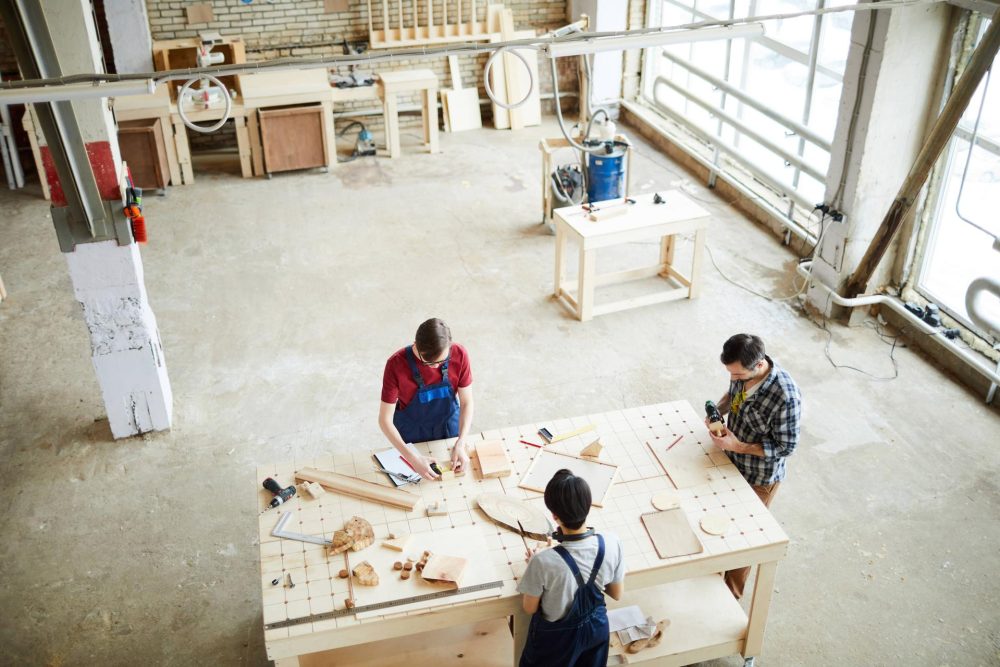 Directly above view of busy carpentry workers standing at wooden workbench and assembling interior details and toys in modern workshop